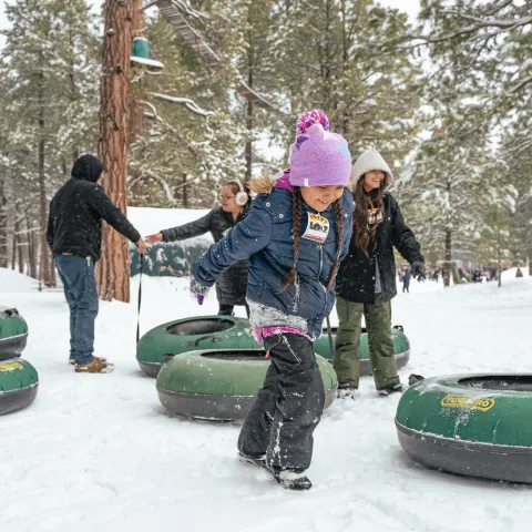 Kids play with snow tubes in a snowy forest, wearing winter clothes and colorful hats.