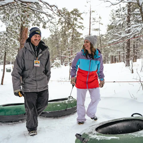 Two people in winter clothes smile while pulling snow tubes in a snowy forest.