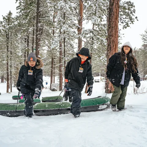 Three children in winter clothes pulling snow tubes through a snowy forest.