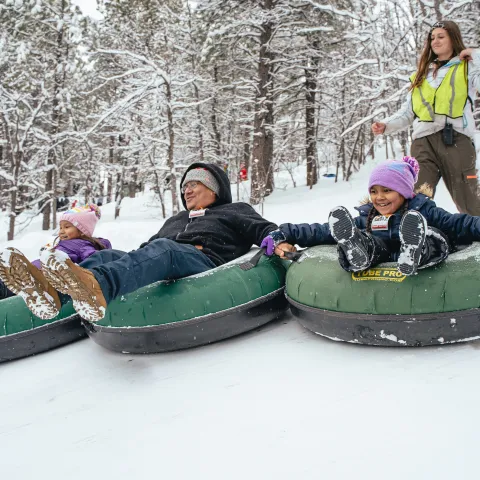 Three people snow tubing down a snowy hill, watched by a woman in a safety vest.