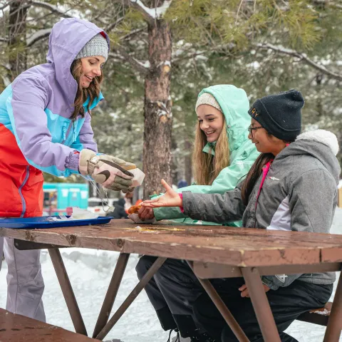 Three people in winter clothing sharing food at a snowy picnic table.
