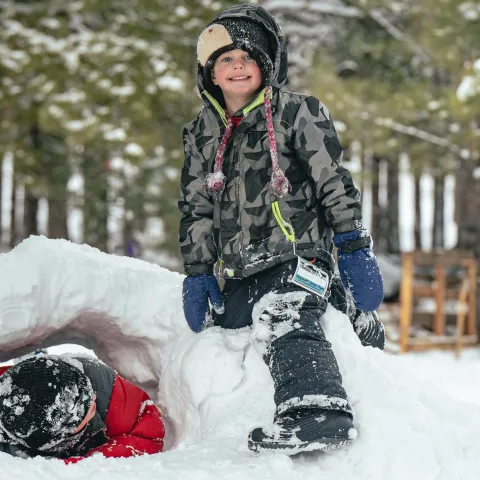 Two children playing in snow, one sitting on a snow tunnel, surrounded by snowy trees.