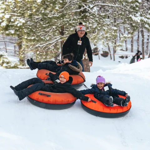 Three kids snow tubing down a hill with an adult standing behind in a snowy forest.