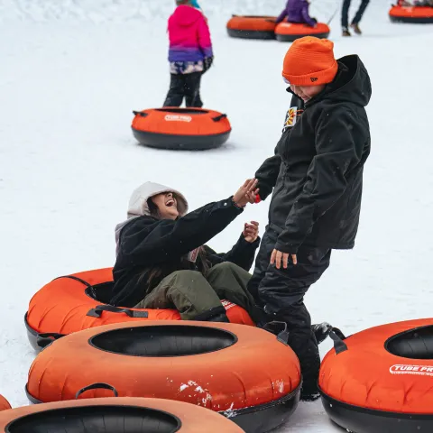 Two people laughing in snow with orange inflatable tubes, dressed in winter clothes.