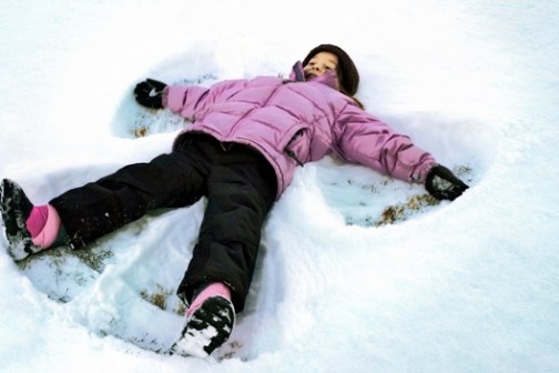 Child in pink jacket making a snow angel on snowy ground.